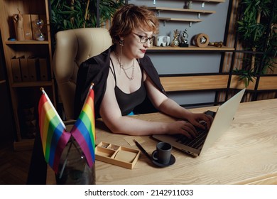 Lady Boss With Short Red Hair Sitting At The Table With Lgbt Flag Working On Laptop Against The Office Background.