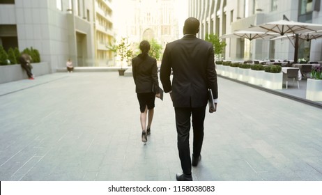 Lady Boss Walking By Office Building Followed By Male Bodyguard, Back View