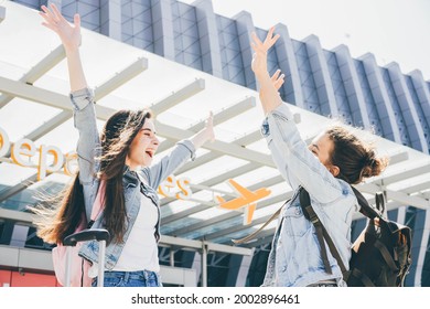 Lady Brunette In Denim Jacket Meets Approaching Girl Friend With Large Black Bag And Greets Hugging Against Local Airport Terminal Close View. Woman Happy To See Each Other. Excited And Amazed