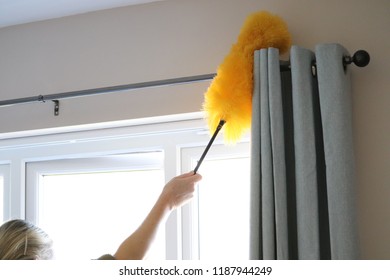 Lady Cleaning Around Window And Curtain Pole With Bright Yellow Fluffy Duster On A Stick