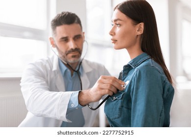 Lady coming to clinic for heart and lungs checkup, male doctor using stethoscope, listening to female patient's breath or heartbeat, sitting in clinic office - Shutterstock ID 2277752915