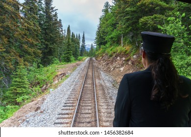 Lady Conductor Is Looking From The Train. Skagway, Alaska She Is Watching Railways And Forest. She Is Wearing Black Uniform And Professional Hat. Dark Hair Woman Is Working On A Rail Road.