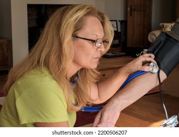 Lady Doctor Mid 50's, Uses A Stethoscope To Listen For The Heart Beat, Whilst Taking A Patients Blood Pressure At Home. Patient Interaction On A Home Visit. 
