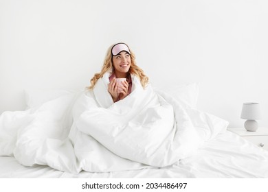 Lady enjoy good morning, weekend and vacation, delicious coffee at free time after great sleep. Satisfied pretty young caucasian woman in sleep mask wrapped in blanket sits on white bed in bedroom - Powered by Shutterstock