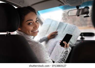 Lady enjoying comfortable auto journey sitting on driver seat. Happy cute millennial african american lady holding map and smartphone with blank screen, turns and looks at passenger from behind
