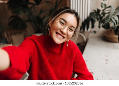 Lady in glasses and massive earrings makes selfie. Asian girl dressed in red knitted sweater posing against background of plants in room - Shutterstock ID 1385232536