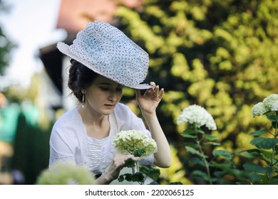 A Lady In A Hat In The Fashion Of The Early 20th Century Walks In A Garden. Portrait Of A Young Slender Woman In A 1910s Edvardian Costume. 