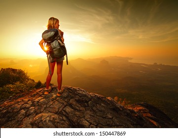 Lady hiker with backpack standing on top of a mountain and enjoying sunrise