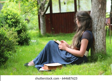 Lady in long blue dress with long hair sitting on the green grass reading the book. The girl sitting under the tree and writing the letter