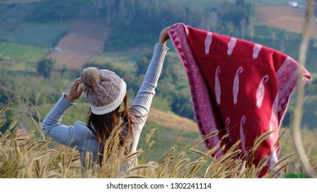 A Lady In Long Sleeves Sweater And Knitting Hat Wave Up The Red Pashmina Scarf Among Grass Field And Blur Mountain Background