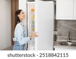 Lady looking inside of fridge while standing in kitchen, opening refrigerator and searching for food, rear view with copy space