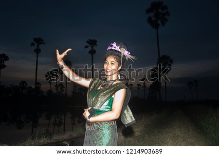 The lady in Middle thai classical dancing suit is posing pattern of traditional dancing,and sugar palm in background at twilight. 