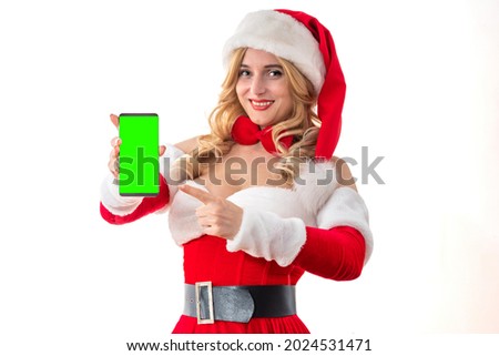 Lady Mrs. Santa Claus smiling happily pointing index finger to smartphone, copy space, green screen mockup. Pretty woman in red dress, white fur. Cheerful girl in Santa’s hat. Christmas party booking