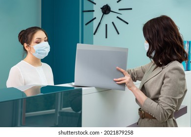 Lady Office Manager With Protective Mask Shows Registration Book To Woman Client Standing At Reception In Contemporary Hospital Close Side View