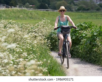 A lady on her electric bike cycles on a cycle route path between white wild flowers.She has handlebar bag and wears a straw hat and sunglasses.