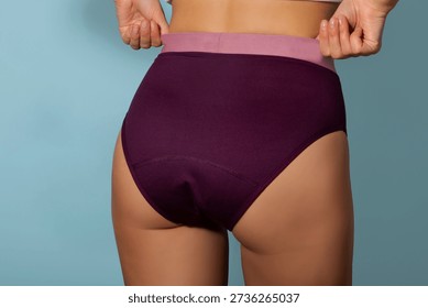 Lady pulling up period underwear while standing against blue studio background, using menstrual panties for comfort