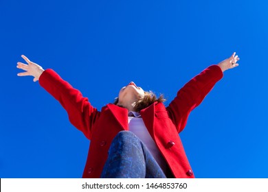 Lady raises her arms and looks to sky. An elated Caucasian woman, wearing a red coat, has her arms in the air with joy on a bright winter's day against a blue sky, shot from a low angle perspective. - Shutterstock ID 1464853760