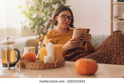 Lady reads a book in blanket on the sofa in front of teapot and candles. - Shutterstock ID 2201515541