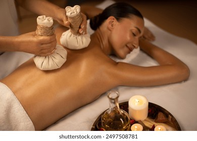 Lady receiving hot herbal ball spa massage body treatment, masseur gently compresses herb bags on woman back in warm lighting of candles at spa salon - Powered by Shutterstock