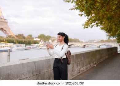 Lady Standing On Embankment Not Far From Eiffel Tower In Paris And Watching Pictures Of Landmarks Made During Day On Mobile. Girl Came To France To Visit Pen Pal And Decided To Go On City Tour. Pretty