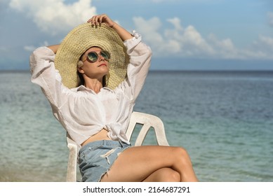 A Lady In A Straw Hat, Sunglasses, White Shirt And Denim Shorts Is Sitting On A Chair. A Young Girl Is Relaxing On The Beach. Travel, Happy Holidays, Tourism, Philippines, Summer Season, Copy Space