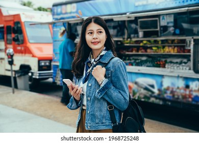 lady studying abroad with smartphone in hand is looking for food truck special recommended online. portrait girl looking away with smile is standing in front of vendor trailers at lunchtime - Powered by Shutterstock