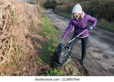 A Lady Uses A Litter Pick Up Tool To Collect A Plastic Bottle And Place It In A Black Bin Bag.She Wears Plastic Gloves And Stands On Verge Of A Rural Country Lane.Pollution.Garbage.
