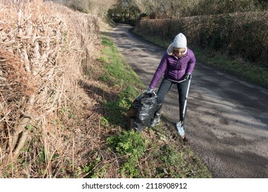 A Lady Uses A Litter Pick Up Tool To Collect A Plastic Bottle And Place It In A Black Bin Bag.She Wears Plastic Gloves And Stands On Verge Of A Rural Country Lane.Pollution.