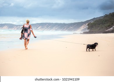 A lady walking her Dachshund dog along a sandy beach with rain clouds in the sky above the sand dunes