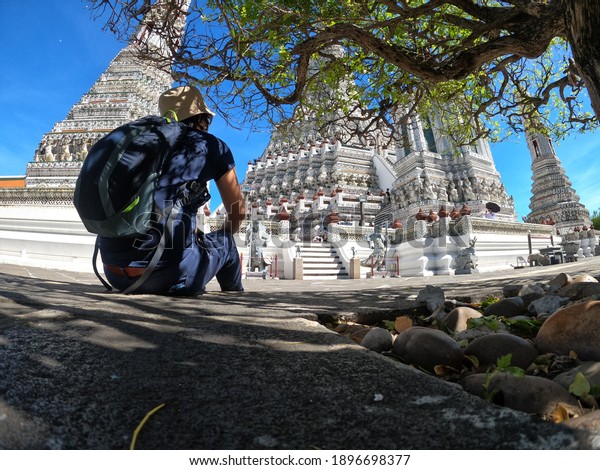 A lady wearing dark blue shirts\
and pants, beige hat sitting in shade looking at the white temple\
in a sunny day, Temple of Dawn, Wat Arun, Bangkok,\
Thailand