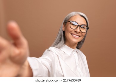 Lady Wearing Glasses Smiling And Looks At The Camera Isolated On Beige. Cheerful Senior Asian Woman Taking Selfie. POV