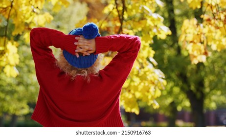 Laid-back Caucasian Young Person Standing Backwards To The Camera In Red Sweater And Blue Winter Hat. Autumn Colors Trees In The Background. High Quality Photo