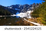 Lake 22 in Granite Falls, WA, with clear, partially frozen waters reflecting rugged cliffs, snow-capped peaks, and pine trees under a bright blue sky. Perfect serene wilderness scene.