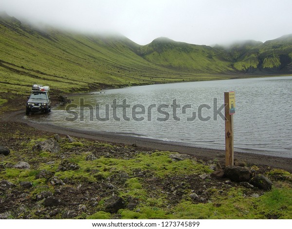 Blautulón lake with car carefully driving around it\
in the water with unusual traffic sign showing right way driving\
through the lake. Lake is enclosed with hills with slopes covered\
with green moss.