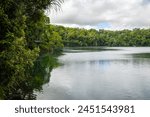 Lake Eacham, a water filled volcanic crater lake surrounded by lush tropical rainforest under an overcast sky on the Atherton Tableland in tropical Queensland, Australia.