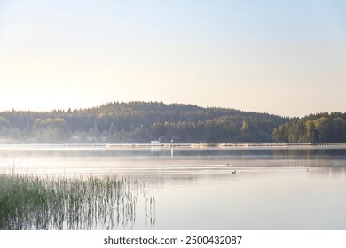 A lake with a foggy morning sky and a small town in the background. The water is calm and the sky is a mix of blue and white - Powered by Shutterstock