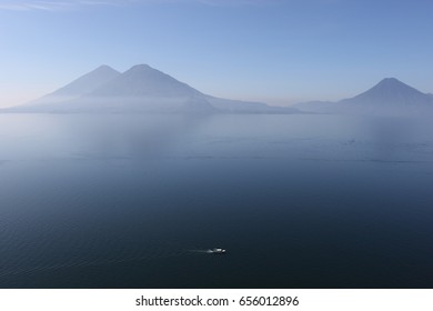 LAKE ATITLÁN - GUATEMALA: Beautiful View Of The Lake From Above, With Three Volcanoes In The Background Mirrored In The Clear Blue Water Where A Boat Passes By Down Below.