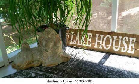 Lake House Outdoor Screened Porch With Pony Tail Palm, Gray Rug, Lake House Sign And Large Painted Vintage Cement Lion. 