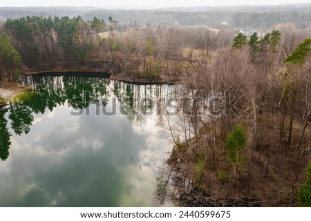 A lake located among forests, whose water is emerald in color. The banks are covered with yellow, dry grasses, leafless trees, between which you can see the green crowns of coniferous trees. The sligh