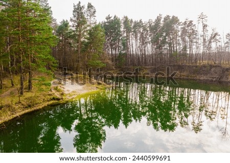 A lake located among forests, whose water is emerald in color. The banks are covered with yellow, dry grasses, leafless trees, between which you can see the green crowns of coniferous trees. The sligh