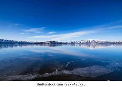 The lake on a clear winter day with fresh blue sky, mirror lake surface, and snowy mountains in the distance. Lake Kussharo in Hokkaido, Japan.