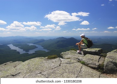 Lake Placid, New York,USA - Jul 16, 2013: young woman with backpack in shorts and tee-shirt sits on cliff edge looking at lakes and mountains panorama. Space for copy. Hiking camping outdoor concept.