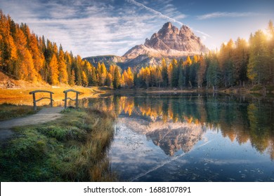 Lake with reflection in mountains at sunrise in autumn in Dolomites, Italy. Landscape with Antorno lake, small wooden bridge, trees with orange leaves, high rocks, blue sky in fall. Colorful forest 