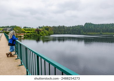 Lake Robertville surrounded by lush pine trees, senior adult woman standing on pedestrian sidewalk admiring landscape, leaning on railing of vehicular bridge, cloudy day in Waimes, Belgium - Powered by Shutterstock