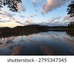 A lake shows clear reflections of clouds and trees as the sun sets. The scene includes a mix of water and land with a few plants along the shore.