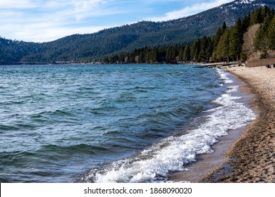 Lake Tahoe During The Day. 

Waves Line The Lakeshore Beach. Sierra Mountains In The Background. 