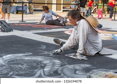 Lake Worth, Florida, USA Fab 23-24, 2019 25Th Annual Street Painting Festival. A Mature Lady Artist Sits Profile On The Asphalt Blends In The Details Of Her Painting With Other Artists Doing The Same.