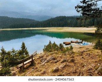 Lakeshore pier with rowing boats and kayaks on mountain lake. Wooden bench, rowboats and canoes on calm water of Black lake surrounded by coniferous forest in foggy morning in Durmitor, Montenegro. - Powered by Shutterstock