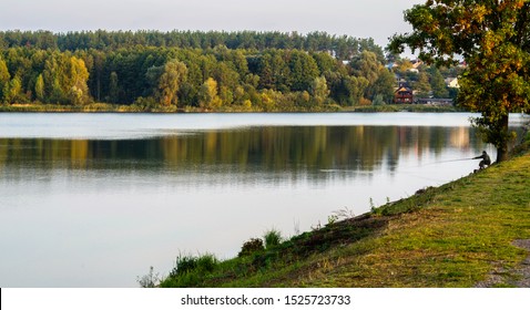 Lakeside with forest and cottages around. Silhouette of a fisherman with a fishing rod.