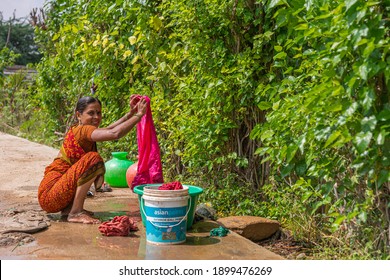 Lakundi, Karnataka, India - November 6, 2013: Squatting Young Woman In Orange Sari Doing Laundry Out Of Buckets Of Water On Street Pavement. Green Foliage As Backdrop.